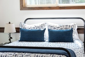 A bed with a patterned bedspread and two blue knitted pillows, a bedside lamp with pine cones, and a window above the bed. - Secluded Lakefront Retreat on 14 Acres with Private Boathouse & Firepit (St. Germain)