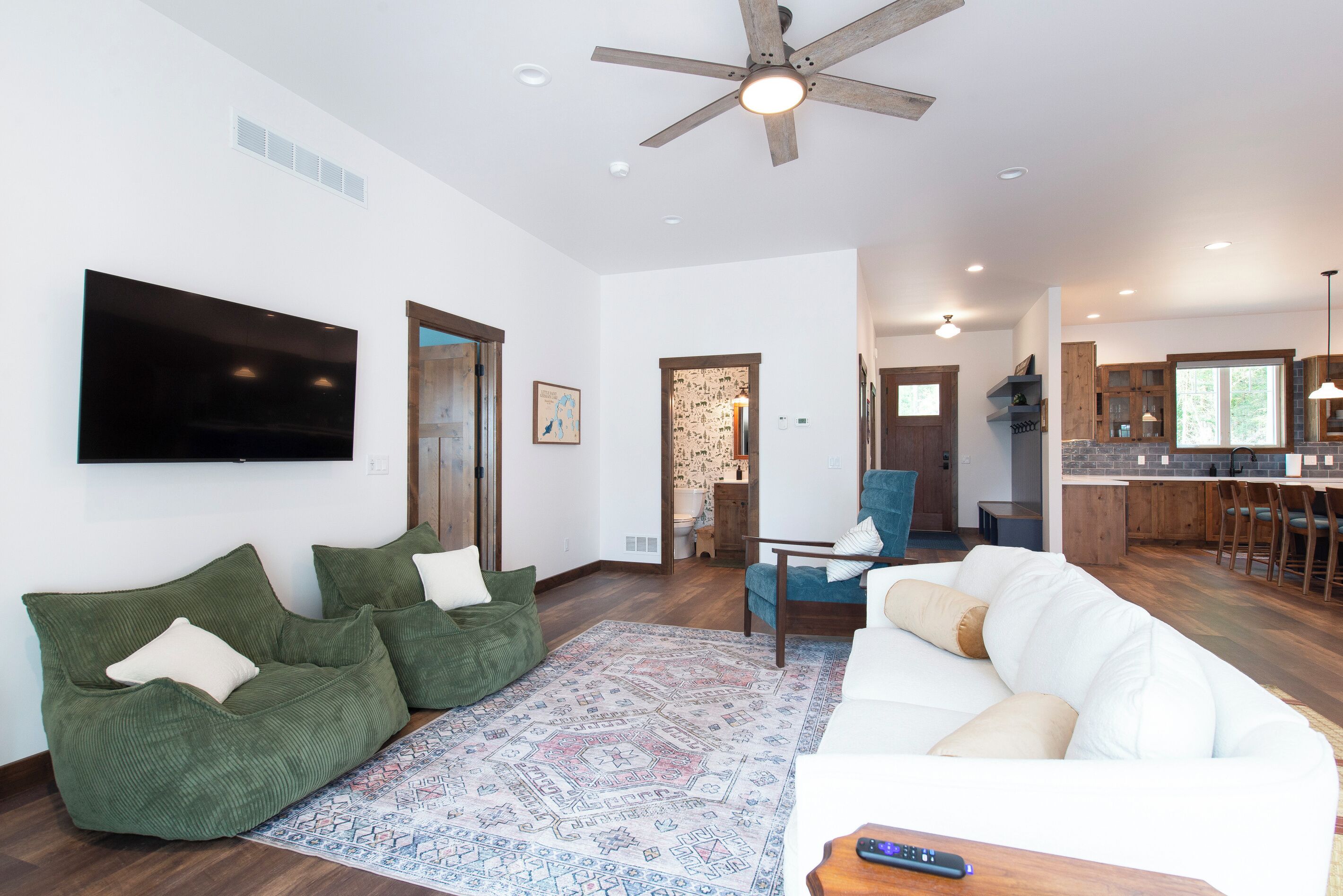 A spacious living area with a large flat-screen TV, green seating, a patterned rug, and a ceiling fan.