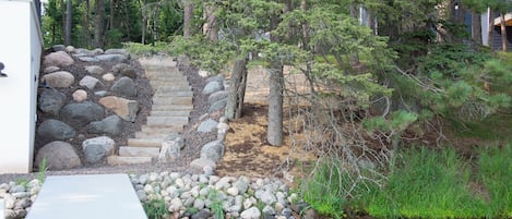 Une terrasse en bois mène à un escalier en pierre, lequel est entouré de rochers et de pins.
