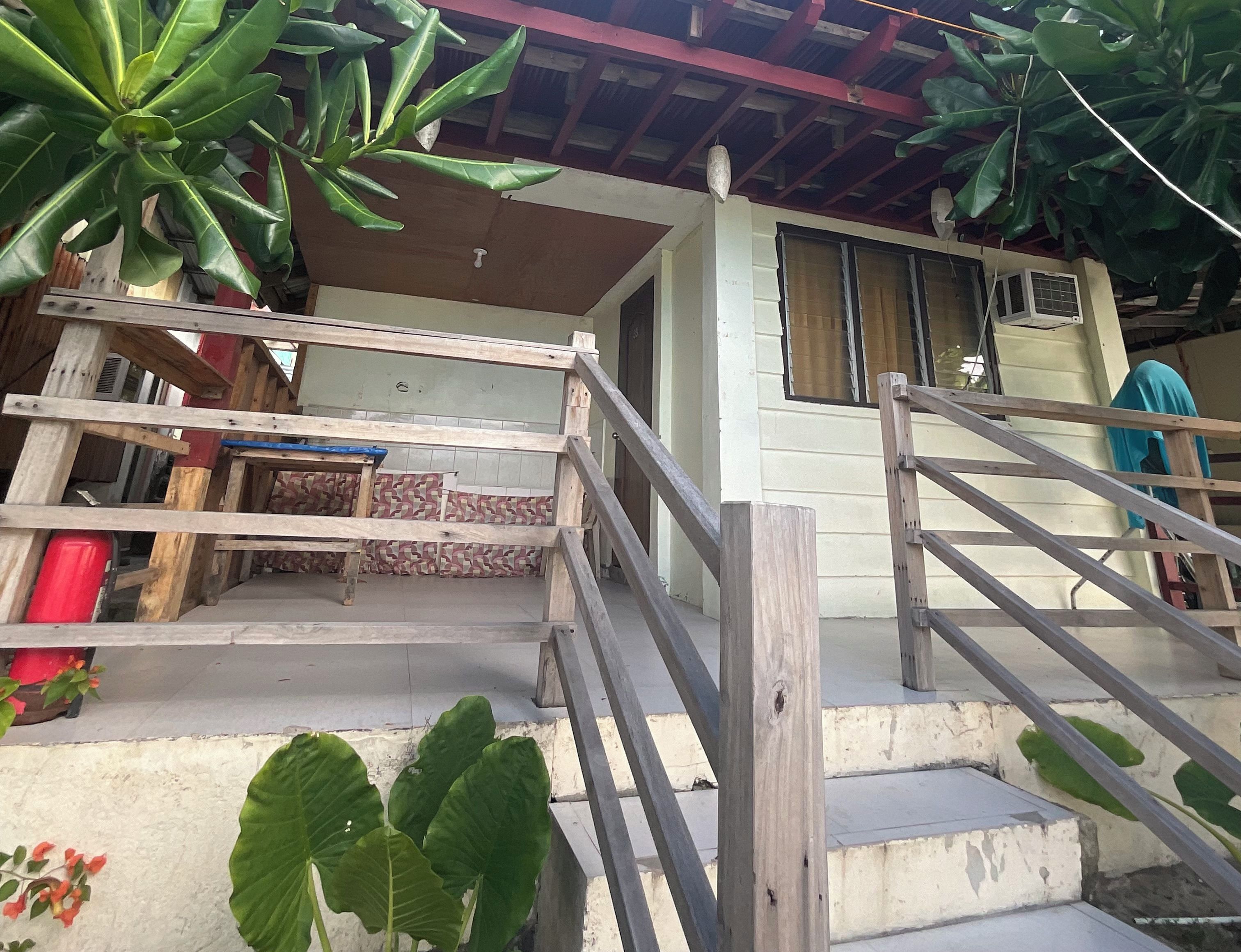 Un bâtiment doté d’un escalier en bois menant à une porte, le tout entouré de grandes plantes vertes.