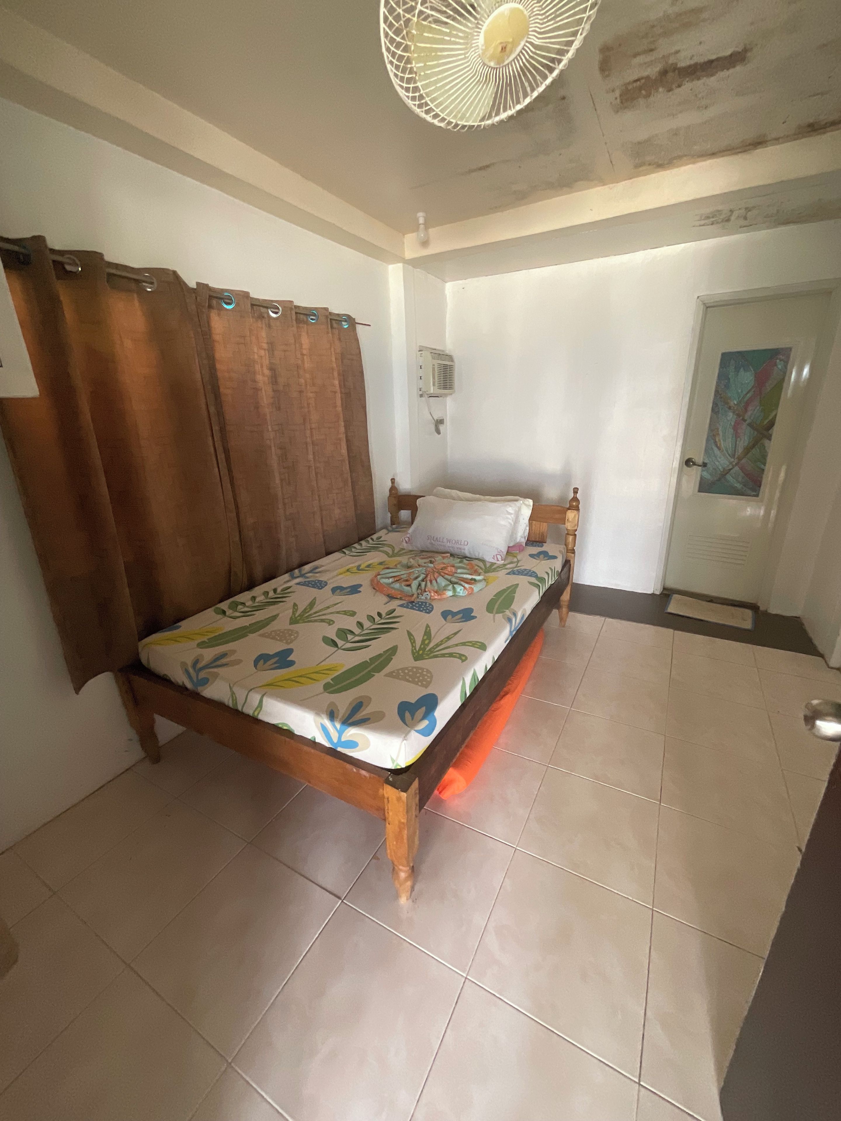 Une chambre à coucher comprenant un lit, des rideaux, un ventilateur de plafond, un climatiseur fixé au mur et une porte ornée d’un tableau.