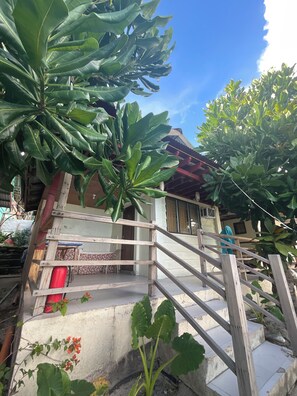 Un escalier en bois, doté d’une rampe, est entouré d’une végétation luxuriante et verdoyante, et mène à un bâtiment dont le toit est recouvert de tuiles.