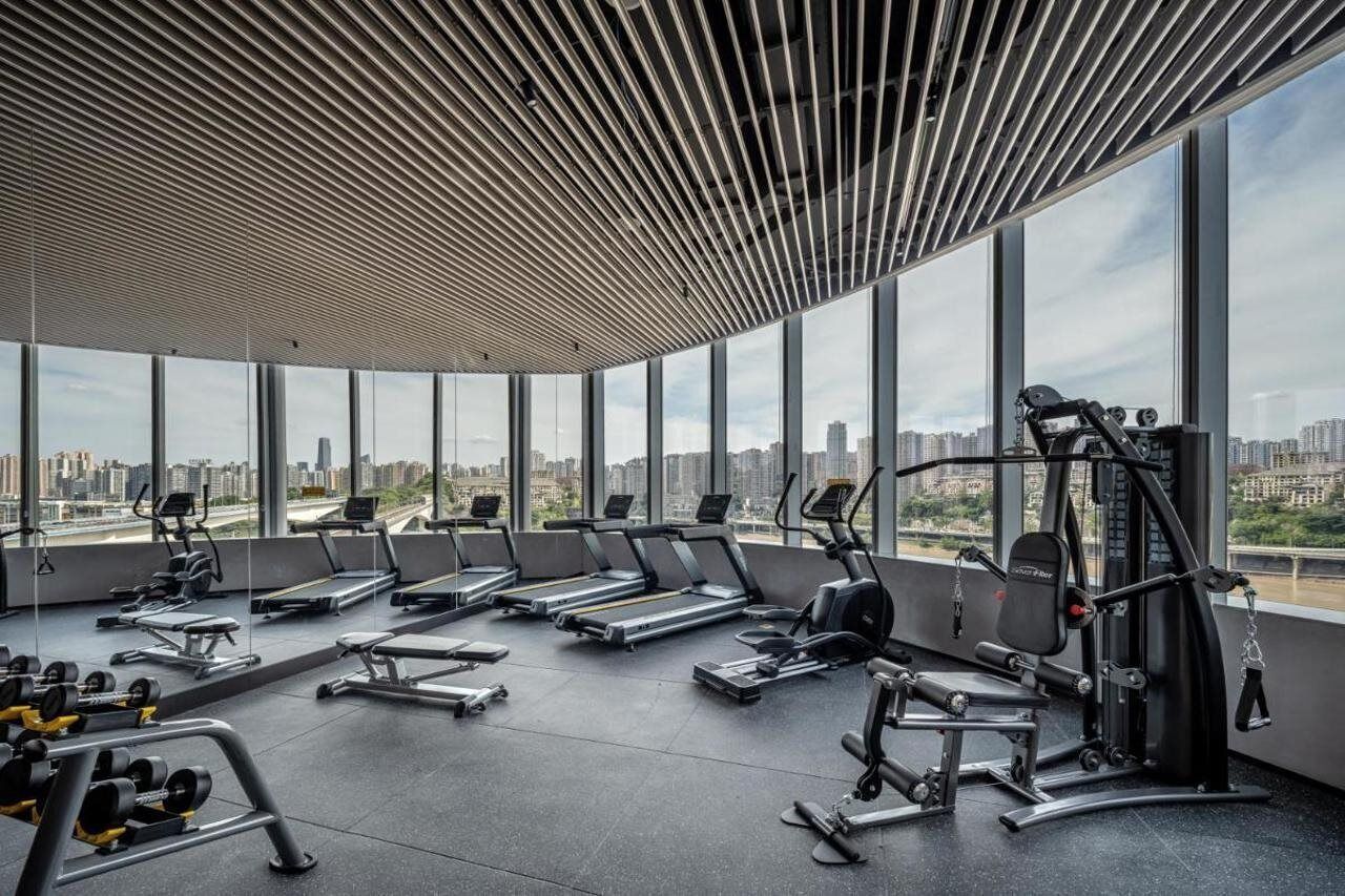 A gym with treadmills, weight machines, and dumbbells, featuring large windows and a city view.