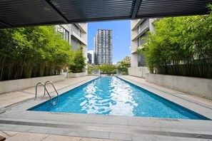 Ein rechteckiger Pool mit einer Metallleiter, umgeben von Grünflächen und modernen Gebäuden im Hintergrund.