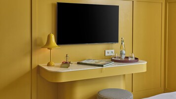 Habitación de hotel con pared amarilla, un televisor de pantalla plana montado sobre una consola blanca, una lámpara de escritorio amarilla, una botella de agua, vasos, un libro y un objeto decorativo pequeño.