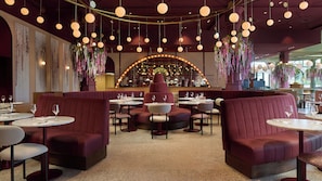 Interior de restaurante con mesas redondas, asientos tapizados en rojo y lámparas colgantes, con adornos florales.