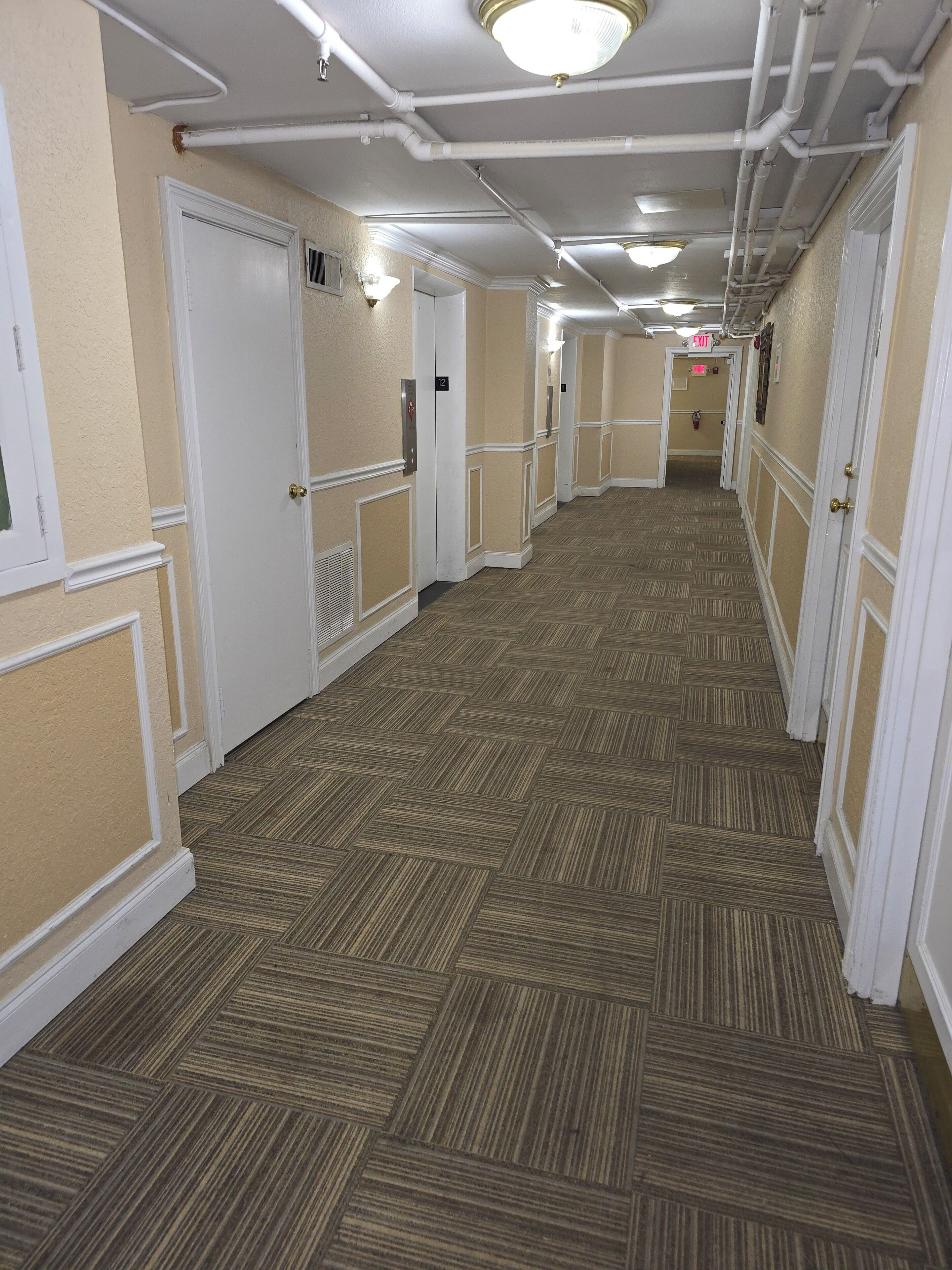 Corredor de hotel com carpete estampado, portas brancas e teto com tubulação aparente.