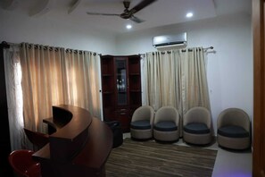 A living room with a curved wooden coffee table, a red chair, and a sofa. There are four beige armchairs, a ceiling fan, and an air conditioning unit.