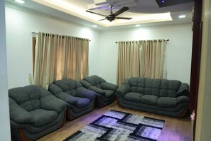 A living room with three grey sofas, beige curtains, a patterned rug, and a ceiling fan.