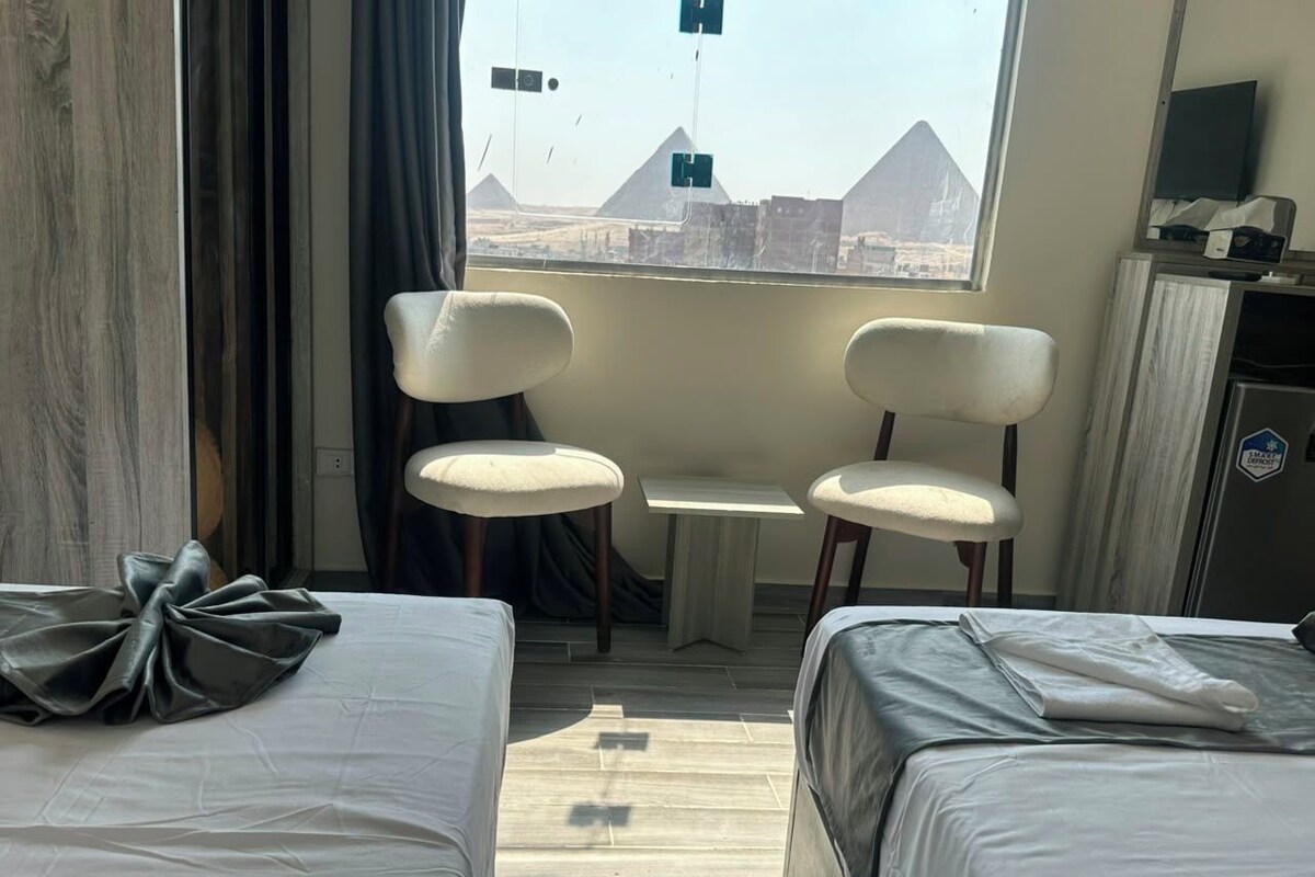 A hotel room with two beds, a small table, two chairs, a TV, and a view of pyramids.