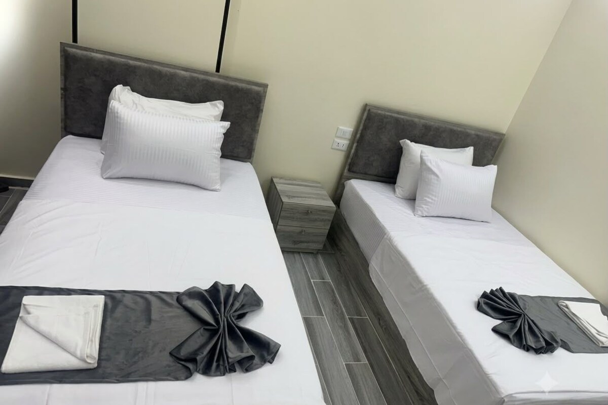 A hotel room with two beds, each with white linens and grey headboards. There is a small bedside table with a lamp and a power outlet on the wall.