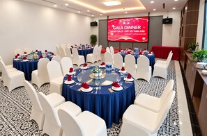 Un salón de conferencias con varias mesas redondas, sillas blancas y una pantalla grande que muestra un evento de cena de gala.