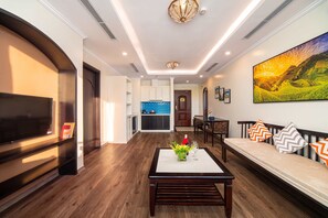 Una sala de estar moderna con un televisor de pantalla plana, una mesa de centro de madera con un florero y una copa de vino, un sofá con cojines estampados y un cuadro de paisaje grande en la pared.