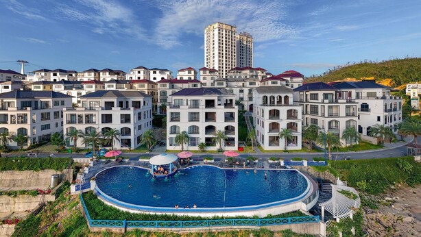 A residential area with a large swimming pool surrounded by modern buildings, a tall hotel in the background, and a hilly landscape. - D'lioro Hotel And Resort (Ha Long)