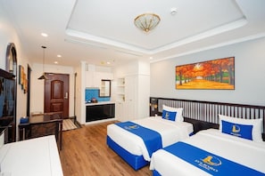 A hotel room with two beds, a desk, a chair, a painting on the wall, and a view of the kitchen.