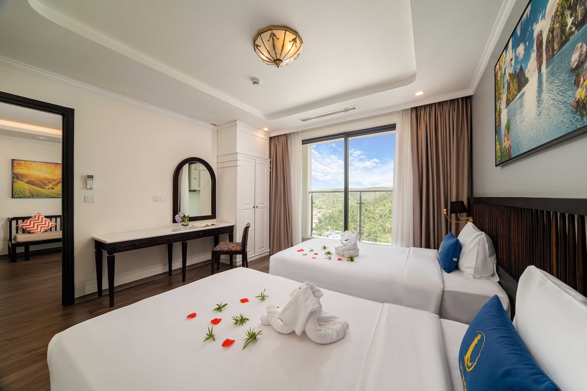 A hotel room with two beds, a desk, a chair, a large window, and a painting on the wall.