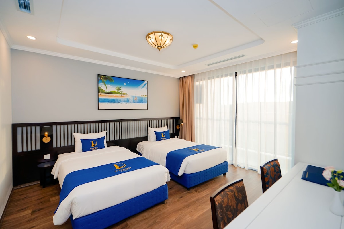 A hotel room with two beds, a painting of a beach scene, a clock, and a window with curtains.