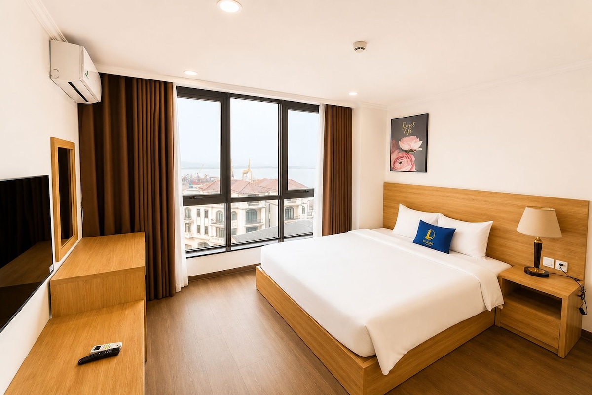A hotel room with a large window, a bed with white bedding, a bedside table with a lamp, and a remote control on the floor.