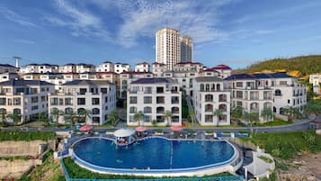 Un quartier résidentiel doté d’une grande piscine entourée de bâtiments modernes, avec un grand hôtel en arrière-plan et un paysage vallonné.