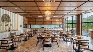 A spacious dining area with multiple tables and chairs, a chandelier, and large windows offering a view of the outdoors.