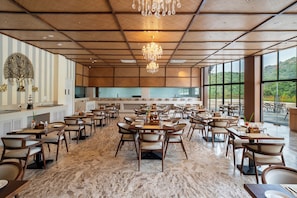 A spacious dining area with multiple tables and chairs, a chandelier, and large windows offering a view of the outdoors.