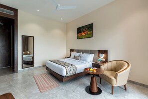 Une chambre d’hôtel avec un lit, une chaise, une table de chevet sur laquelle se trouve un panier de fruits, un tableau au mur et un ventilateur de plafond.