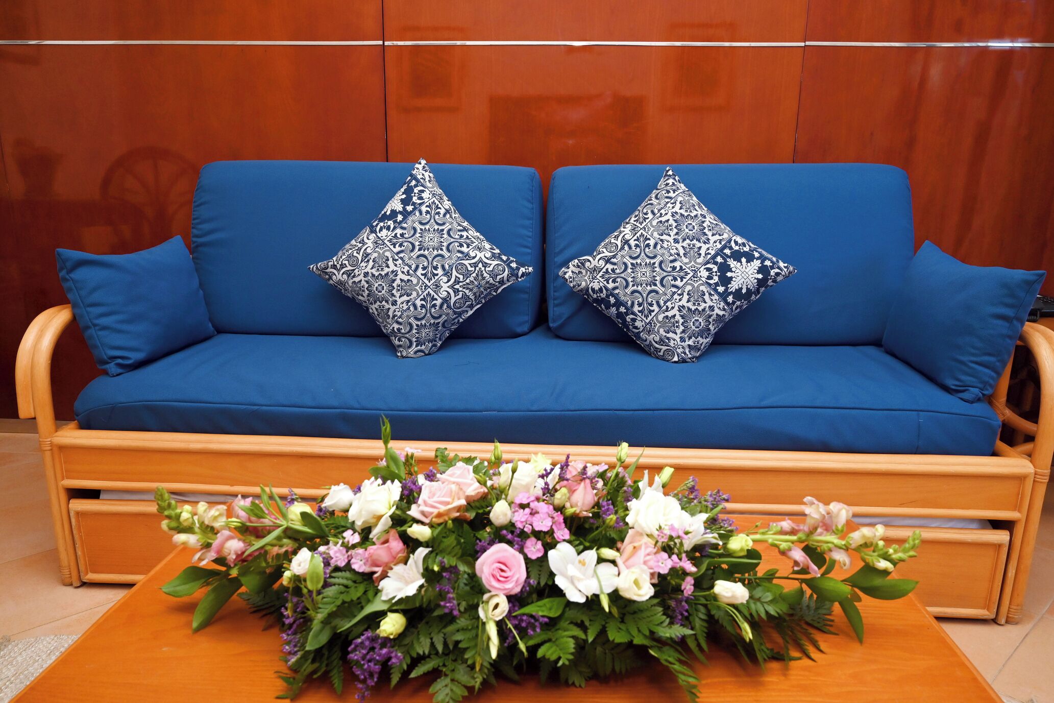 Un sofá azul con cojines estampados, una mesita de madera con un arreglo floral y una pared de madera.