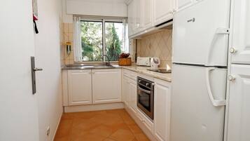 Una cocina compacta con armarios blancos, horno integrado, refrigerador y ventana con vista a árboles.