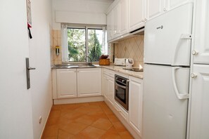 Una cocina compacta con armarios blancos, horno integrado, refrigerador y ventana con vista a árboles.