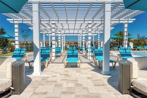 Ein überdachter Außenbereich mit Liegestühlen und Sonnenschirmen, im Hintergrund ein Pool und ein klarer Himmel.
