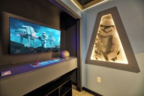 A room with a flat-screen TV mounted on a wall, a gaming console, and a decorative wall piece. - Breezy Palms|Spectacular NEW Villa|NEW WOW Resort (Clermont)
