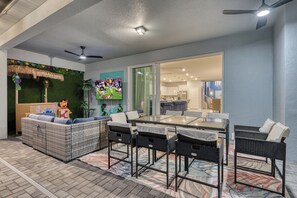 An outdoor patio area with a glass-top table, wicker furniture, and a TV screen displaying a sports game. - Breezy Palms|Spectacular NEW Villa|NEW WOW Resort (Clermont)