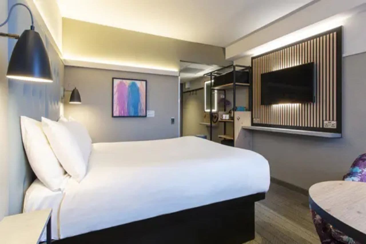 A hotel room with a bed, a desk, a chair, a TV mounted on the wall, and a painting on the wall.