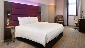 A hotel room with a large bed, a nightstand, a chair, and a desk with a lamp.