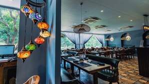 A restaurant interior with hanging decorative lamps, tables set for dining, and large windows offering a view of trees. - Anchaviyo Resort (Vada)