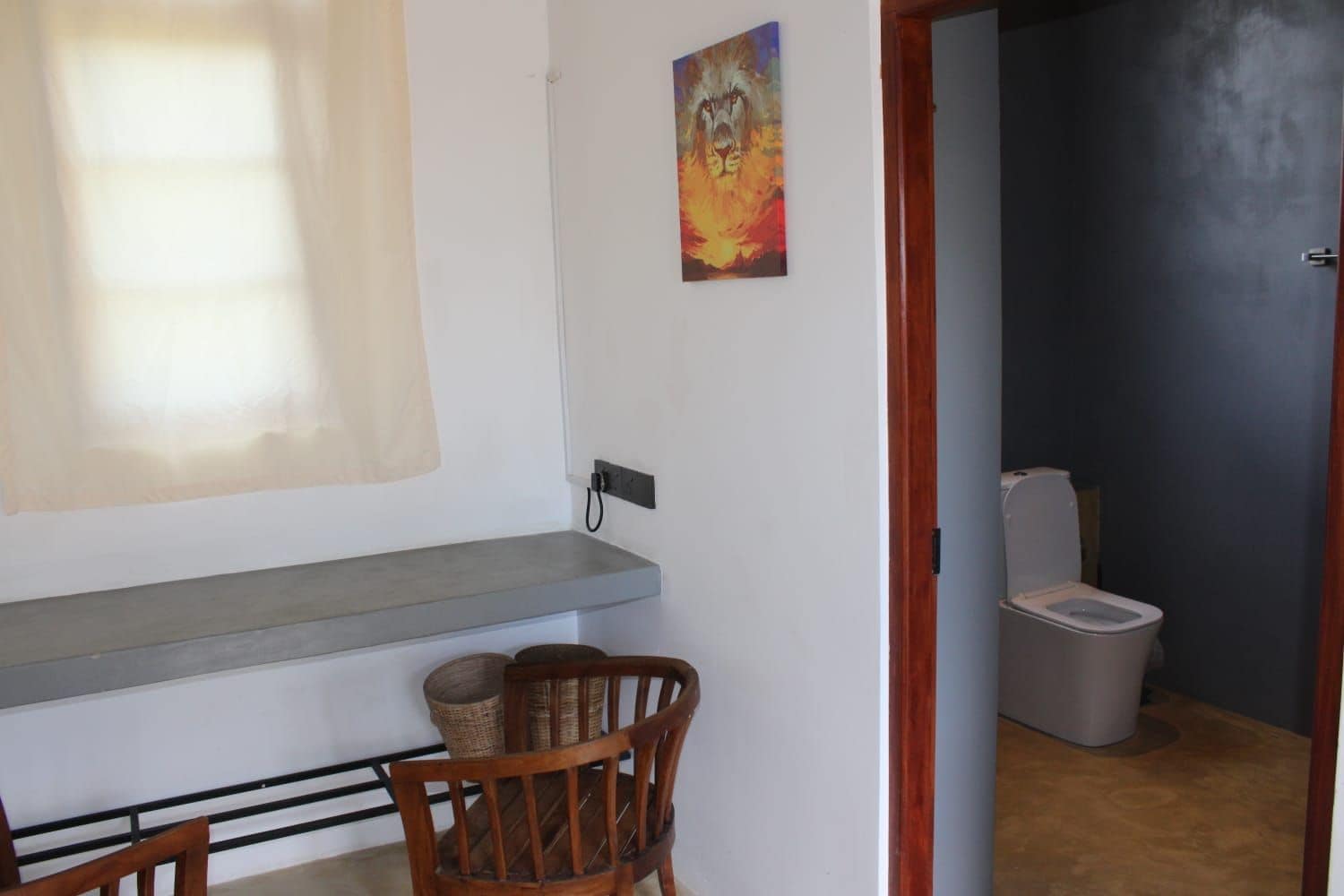 Una habitación con una silla de madera, una estantería, un cuadro y un inodoro visible a través de una puerta abierta.