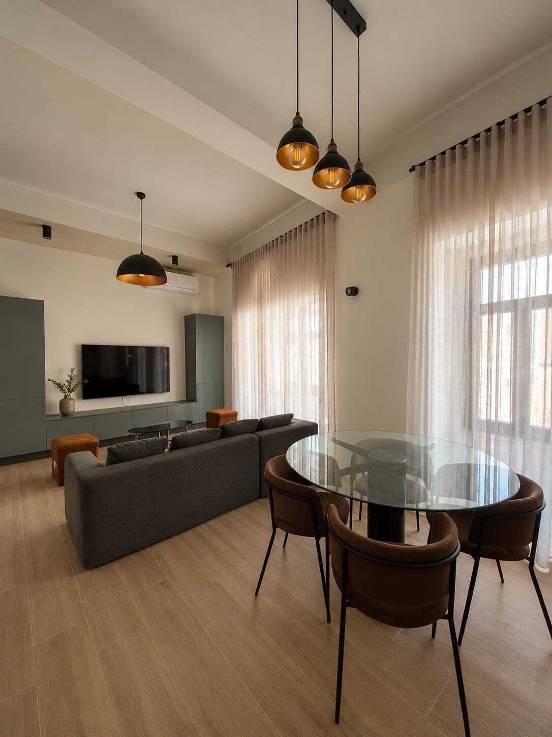 A modern living room with a glass dining table, dark sofas, and pendant lighting.