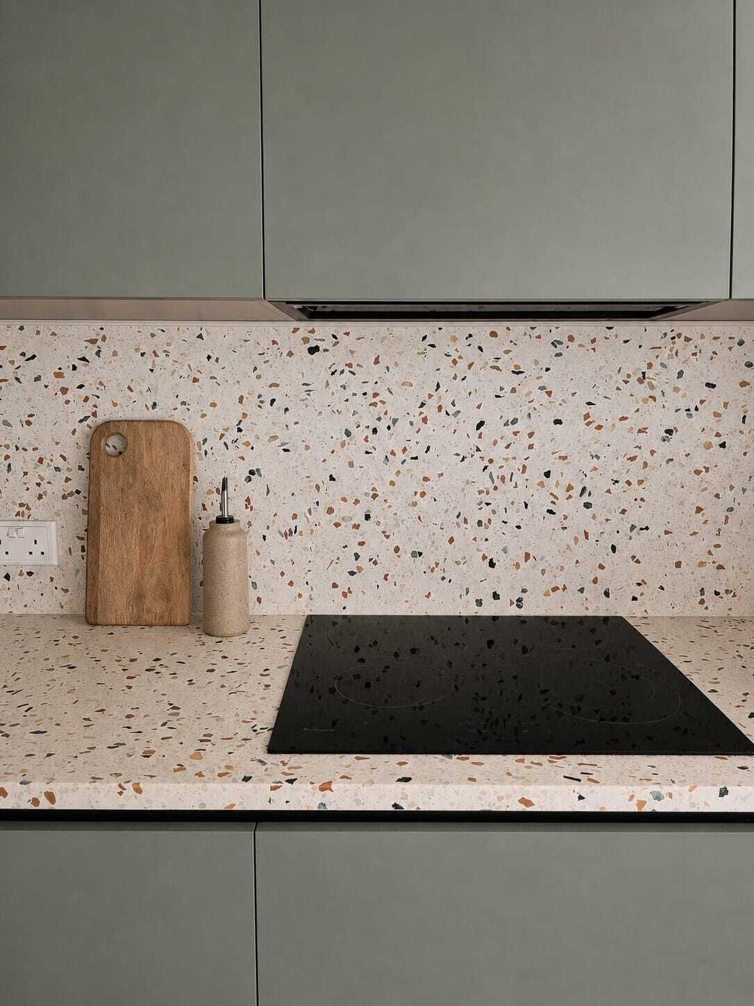 A kitchen with a speckled backsplash, a wooden cutting board, a bottle, and a black countertop.