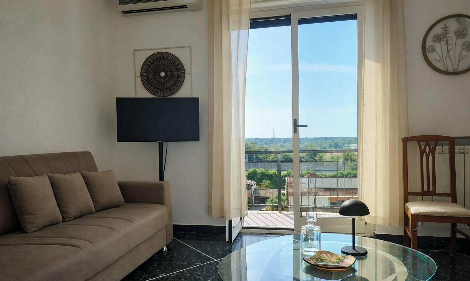 A modern living room with a sofa, a glass table, a chair, a flat-screen TV, and a view of greenery through a sliding door.