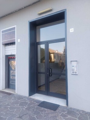 Entrada de un edificio con puerta de vidrio, sistema de teclado para acceder y un pequeño espacio exterior con escalones que llevan a la entrada.