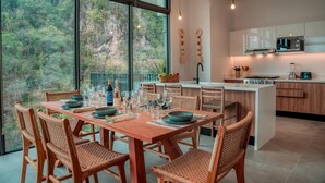 A modern kitchen with a dining area, large windows, and a view of the forest. - Luxury 3BR Oasis | River & Ocean Views 3BD Condo for rent in El Caloso, Puerto v (Puerto Vallarta)