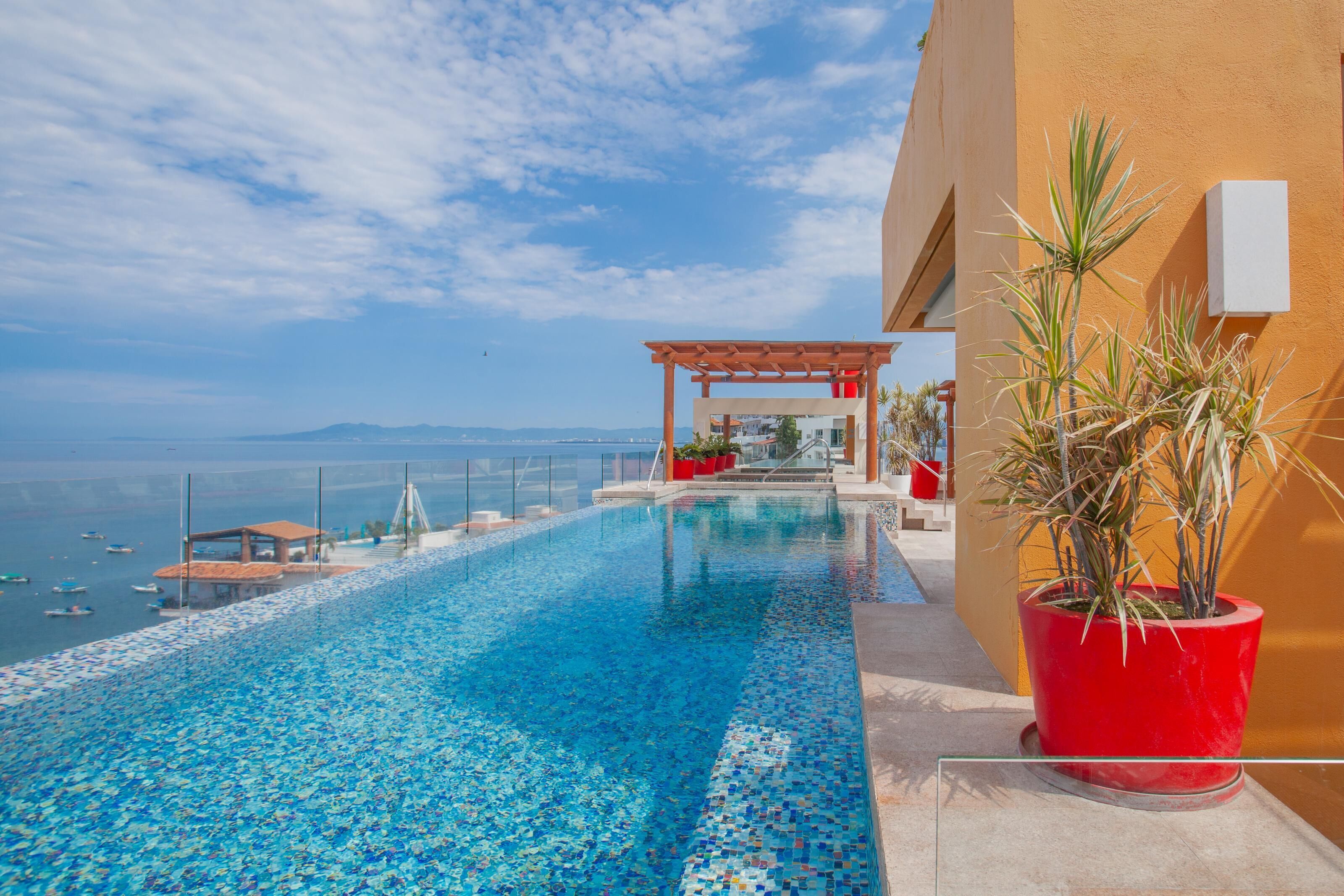 A clear blue infinity pool with a view of the sea and boats, a red potted plant, and a wooden pergola structure.