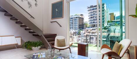 A modern living room with a glass coffee table, a sofa, a chair, a staircase, and a large window offering a city view.