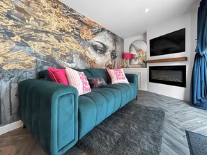 Ein smaragdgrünes Sofa mit vielen Kissen, ein großes abstraktes Gemälde an der Wand, ein Kamin und ein an der Wand befestigter Fernseher.