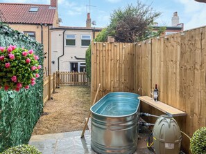 Ein Garten mit Holzzaun, einem Metallwassertank und einer kleinen Blumenrabatte mit rosa Blüten.