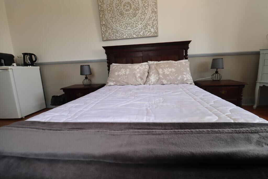 Habitación de hotel con cama, dos mesitas de noche, un tocador, una obra de arte en la pared y un pequeño electrodoméstico blanco.