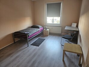 Quarto pequeno e bem iluminado com uma cama de solteiro, uma escrivaninha e uma cadeira.