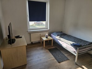 Um quarto pequeno e pouco mobiliado, com uma cama de solteiro, uma escrivaninha com computador, uma mesa pequena e um radiador.