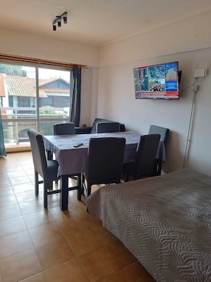 Une pièce comprenant un lit, une table à manger avec des chaises, un téléviseur fixé au mur et une vue sur l’extérieur grâce à une grande fenêtre.
