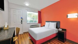 Hotellihuone, jossa on sänky, yöpöytä, tuoli, työpöytä ja ikkuna, jossa on verhot.